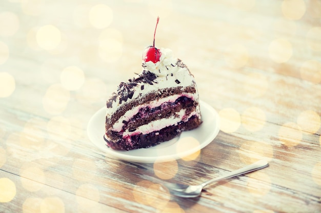 jedzenie, niezdrowe jedzenie, kulinarne, pieczenie i święta koncepcja - kawałek pysznego ciasta z czekoladą wiśniową na spodku z łyżką na drewnianym stole nad świątecznym tłem światła