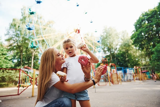 Jedzenie pączków. Wesoła dziewczynka na wrotkach i jej mama wspólnie bawią się w parku przy atrakcjach.