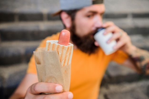 Jedzenie uliczne jest tak dobre. Żywienie miejskiego stylu życia. Beztroski hipster je niezdrowe jedzenie, siedząc na schodach. Przekąska głodny człowiek. Niezdrowe jedzenie. Facet je hot doga. Brodaty mężczyzna ugryźć smaczną kiełbasę i wypić papierowy kubek.