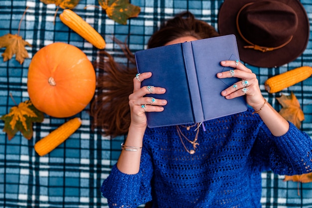 Jesienna kobieta w dzianinowym swetrze z książką leży na niebieskim przytulnym kocu w kratę pokrytą suchymi liśćmi klonu