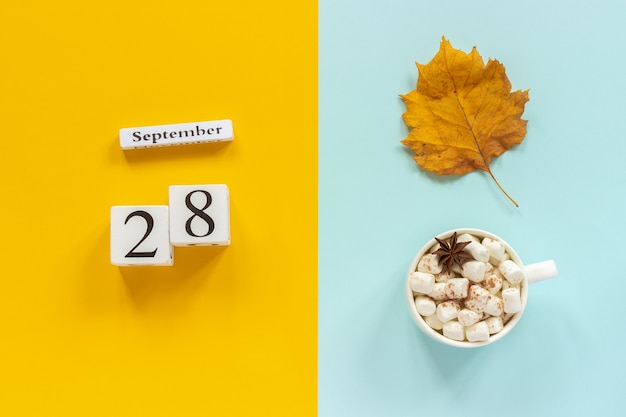 Jesienna kompozycja. Drewniany kalendarz 28 września, filiżanka kakao z piankami i żółtymi jesiennymi liśćmi na żółtym niebieskim tle. Widok z góry Mieszkanie świeckich koncepcja makieta Witam września.