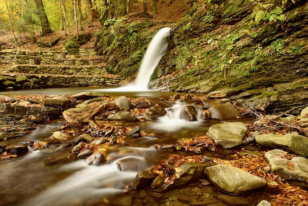 Jesienny górski strumień wodospadu w skałach z kolorowymi czerwonymi opadłymi suchymi liśćmi, naturalne tło sezonowe