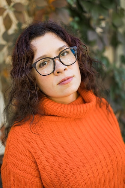 Jesienny portret atrakcyjnej młodej kobiety w stylowych okularach w modnym pomarańczowym swetrze z dzianiny na tle liści Dziewczyna spaceruje po mieście