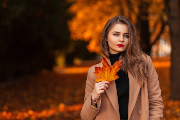 Jesienny portret pięknej młodej dziewczyny z kolorowym czerwono-pomarańczowym liściem klonu w modnym beżowym płaszczu ze swetrem spaceruje po parku z liśćmi. Miejsce na tekst, skopiuj miejsce