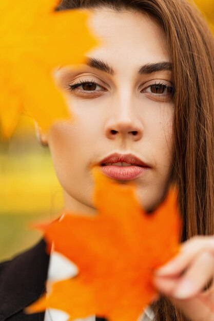 Jesienny portret pięknej mody kobiety z ładną twarzą w eleganckich ubraniach z kolorowymi jasnymi jesiennymi liśćmi spaceruje po jesiennym parku i patrzy w kamerę