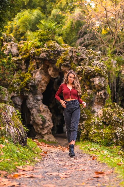 Jesienny styl życia blondynka kaukaska dziewczyna w czerwonym swetrze i dżinsach i na wysokich obcasach, podziwiając przyrodę w parku z naturalną jaskinią