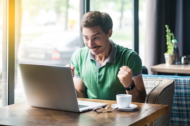 Jestem zwycięzcą Młody szczęśliwy zaskoczony biznesmen w zielonej koszulce siedzący patrząc na ekran laptopa i cieszący się z jego sukcesu w biznesie i koncepcji freelancingu strzał w pobliżu dużego okna w ciągu dnia