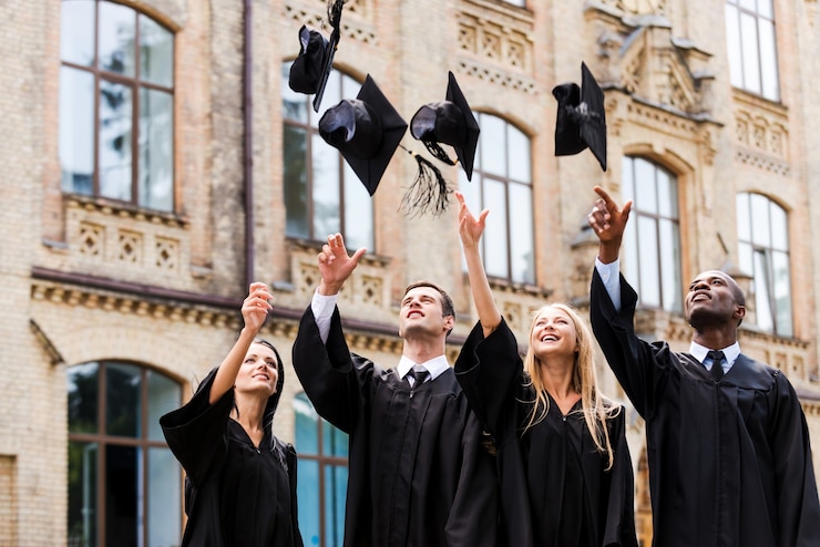 Zdjęcie jesteśmy wreszcie absolwentami! czterech szczęśliwych absolwentów college'ów w sukniach dyplomowych rzucających deskami z zaprawy murarskiej i uśmiechających się, stojąc w pobliżu uniwersytetu