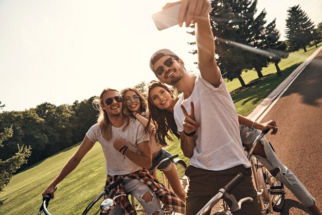 Jeszcze tylko jeden strzał. Grupa szczęśliwych młodych ludzi w strojach codziennych, biorących selfie i uśmiechających się podczas wspólnej jazdy na rowerze na świeżym powietrzu