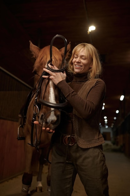 Jeździec kobieta zaprzęgająca konia w stajni klubu jeździeckiego lub rancza. Kobieta amazonka oddanie uprzęży na twarz ogiera widok z przodu strzał