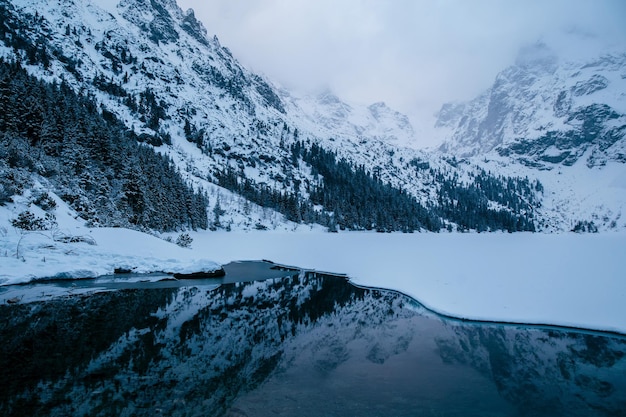 Jezioro Morskie Oko znajduje się przed majestatycznym śnieżnym łańcuchem górskim, tworząc oszałamiający krajobraz naturalny. Niebo jest wypełnione puszystymi chmurami, wzmacniającymi atmosferę Tatrów Polska