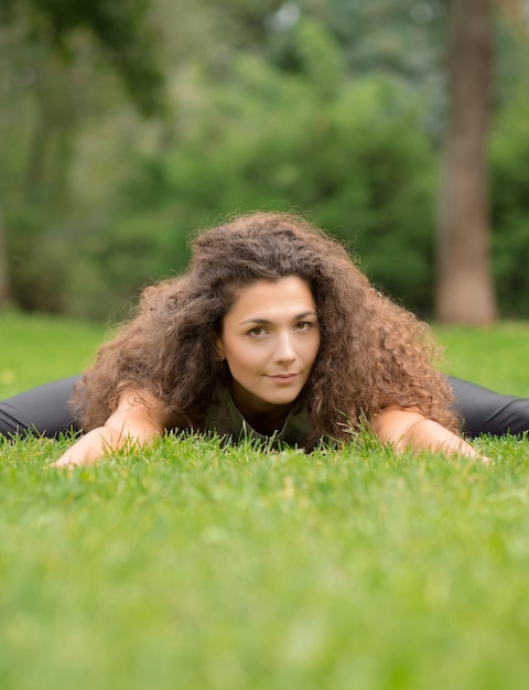 Joga lub fitness w letnim parku. Widok z przodu młodej kobiety z długimi kręconymi włosami leżącej na zielonej trawie