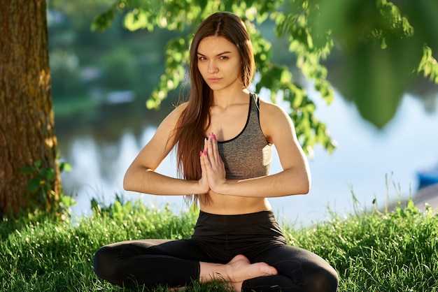 Joga w parku z widokiem na rzekę, w słońcu. Młoda kobieta w pozycji lotosu, siedząc na zielonej trawie. Pojęcie spokoju i medytacji.