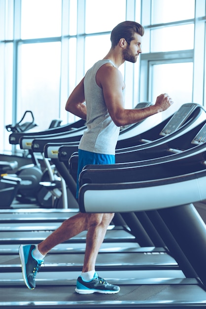 Jogging na bieżni. Widok z boku na całej długości młodego mężczyzny w odzieży sportowej biegającej na bieżni w siłowni