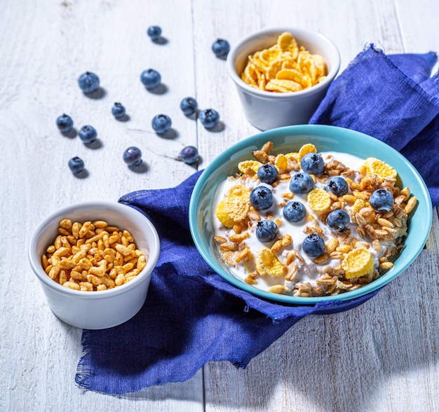 Jogurt, miseczki z muesli, jagodami, płatkami kukurydzianymi i dmuchanym ryżem na białej powierzchni. Zdrowe śniadanie