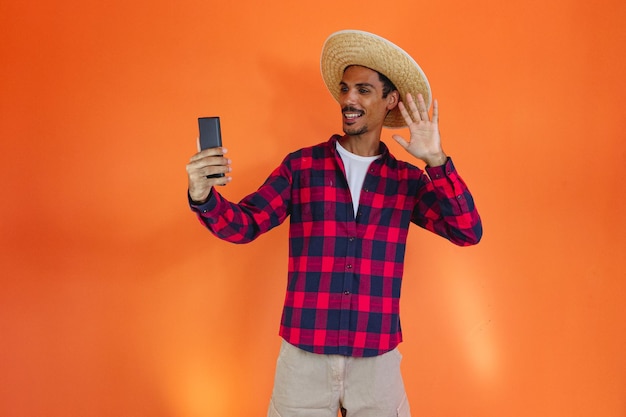 Junina Party Black Man With Outfit trzymając telefon komórkowy izolowany na pomarańczowym tle Młody mężczyzna ubrany w tradycyjne stroje na Festa Junina brazylijski czerwcowy festiwal