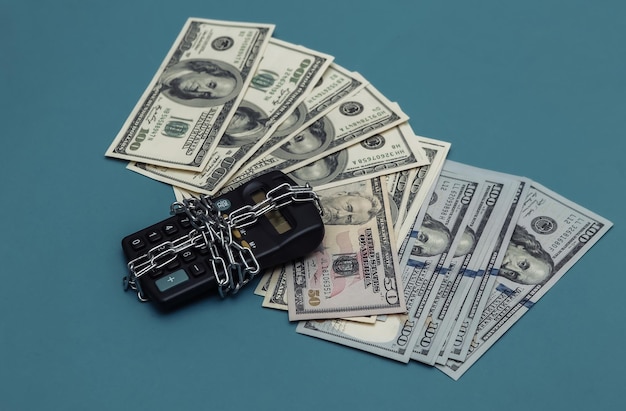 Kalkulator zawinięty w stalowy łańcuch i studolarowe banknoty na niebieskim tle