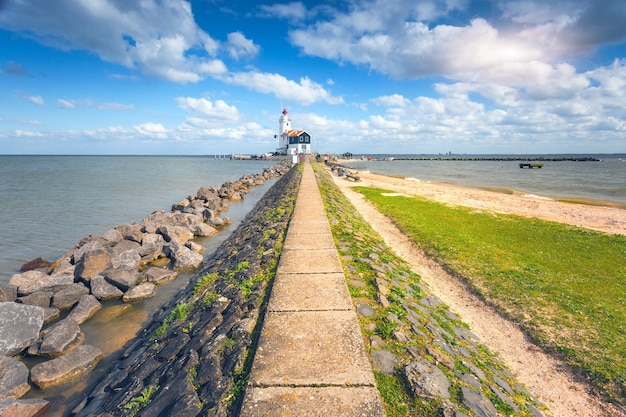 Kamienna ścieżka prowadząca do latarni morskiej na wybrzeżu morza na tle błękitnego nieba z chmurami o zachodzie słońca wiosną w Holandii. Krajobraz ze szlakiem, kamieniami, trawą, plażą, oceanem i latarnią morską. Natura