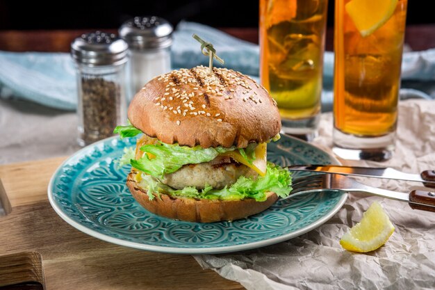 Kanapka z burgerem z kurczaka, serem i sałatą. Burger leży na stole z papierem i solniczkami i pieprzniczkami. Oprócz dwóch koktajli log Island Iced