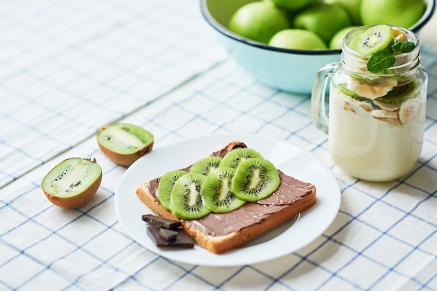 Kanapka z masłem orzechowym i kiwi, zielonymi jabłkami, jogurtem z bananem i kiwi, ciekawy pomysł na śniadanie dla dzieci, deser lub posiłek świąteczny, widok z góry leżał płasko, zdrowe jedzenie dla dzieci. Dzień dobry