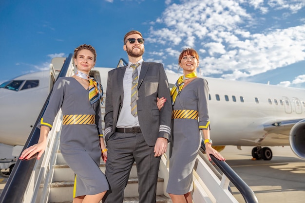 Kapitan lotnictwa i dwie stewardessy stoją na schodach samolotu i uśmiechają się