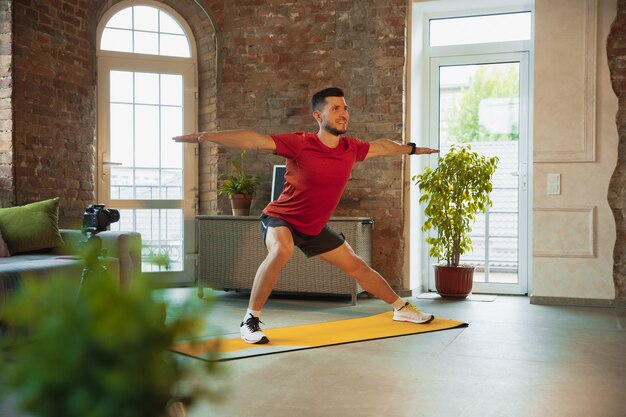 Kardio. Młody kaukaski mężczyzna trenuje w domu podczas kwarantanny epidemii koronawirusa, robi ćwiczenia fitness, aerobik. Nagrywanie wideo lub przesyłanie strumieniowe online. Wellness, sport, koncepcja ruchu.