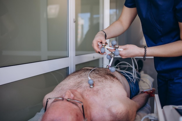 Kardiolog badający starszego pacjenta za pomocą urządzenia Holtera na oddziale kardiologii w szpitalu Kobieta lekarz dołącza czujniki do pomiaru EKG i ciśnienia krwi Dojrzały mężczyzna na kanapie kliniki