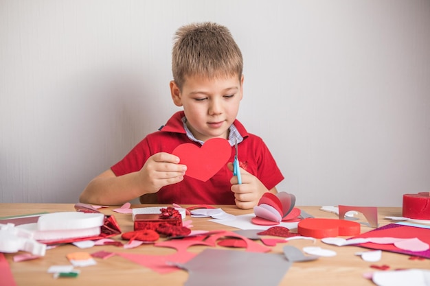 Kartka świąteczna DIY z czerwonym papierowym sercem, symbolem miłości. Chłopiec dziecko sprawia, że dzień matki, Walentynki, kartkę z życzeniami. Hobby, koncepcja sztuki dla dzieci, prezent własnymi rękami, pomysły na majsterkowanie dla dzieci