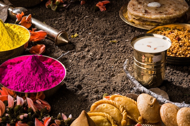 Kartka z życzeniami Happy Holi przedstawiająca tradycyjne indyjskie słodkie i słone potrawy, kwiaty i kolory proszku ułożone na białym tle lub glinie. Selektywne skupienie