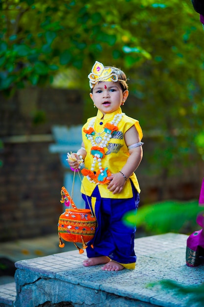 Kartka z życzeniami Happy Janmashtami przedstawiająca małego indyjskiego chłopca udającego Shri Krishna lub kanha/kanhaiya z obrazkiem Dahi Handi i kolorowymi kwiatami.