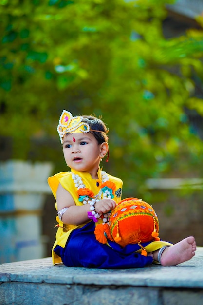 Kartka z życzeniami Happy Janmashtami przedstawiająca małego indyjskiego chłopca udającego Shri Krishna lub kanha/kanhaiya z obrazkiem Dahi Handi i kolorowymi kwiatami.
