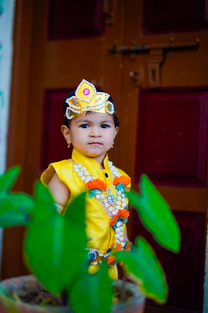 Kartka Z życzeniami Happy Janmashtami Przedstawiająca Małego Indyjskiego Chłopca Udającego Shri Krishna Lub Kanha/kanhaiya Z Obrazkiem Dahi Handi I Kolorowymi Kwiatami.