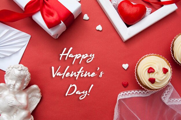 Kartkę z życzeniami z tekstem Happy Valentines Day. Prezent, babeczki, serce, koperta, anioł, wstążki - płaskie walentynki leżały na czerwonym tle.