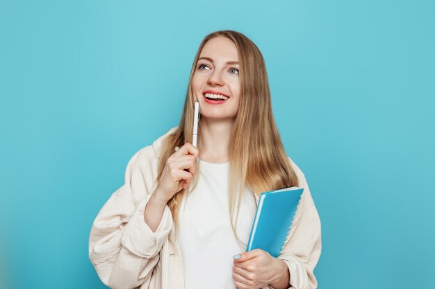 Kaukaska blondynka studentka myśli i marzenia, trzyma notatnik, notatnik, książkę na białym tle na niebieskiej ścianie w studio. Testy, egzaminy, koncepcja edukacji. Skopiuj miejsce