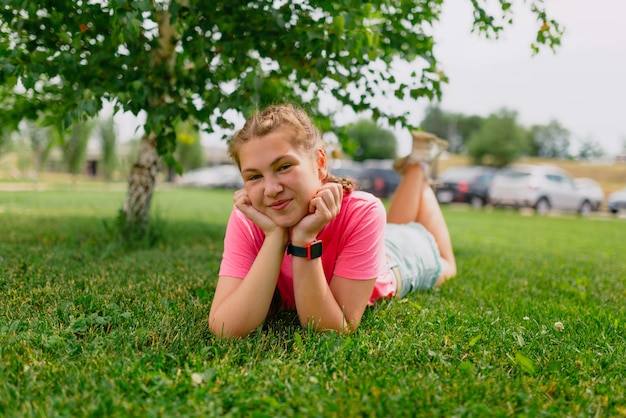 Kaukaska dziewczyna z piegami iw różowej koszulce leży na trawie. Student dziewczyna uśmiecha się i cieszy się piękną letnią pogodą.