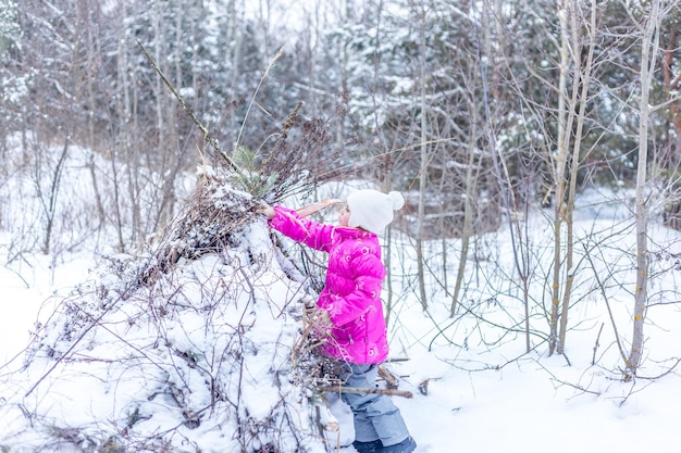 Kaukaska dziewczynka od 5 lat buduje chatę z gałęzi iglastych w zimowym lesie, bawiąc się zimą na świeżym powietrzu
