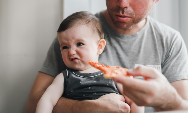 Kaukaska dziewczynka siedzi w ramionach swojego taty, który daje jej kawałek domowej pizzy