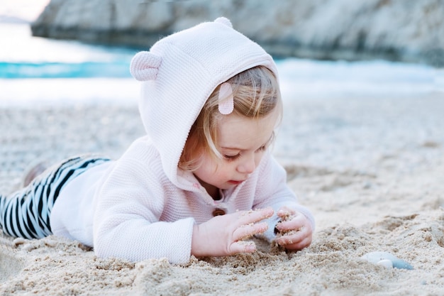 Kaukaska dziewczynka ubrana w koszulę z zabawnymi zwierzęcymi uszami na piaszczystej plaży bawi się kamieniamisiedząc piasek nad błękitnym morzem