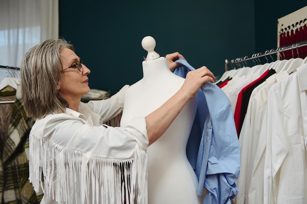 Kaukaska kobieta, projektantka mody, krawcowa, przymierza niebieską koszulę z nowej kolekcji na manekinie w swoim warsztacie projektowania odzieży i krawiectwa