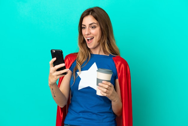 Kaukaska kobieta Super Hero na białym tle na niebieskim tle trzymająca kawę na wynos i telefon komórkowy