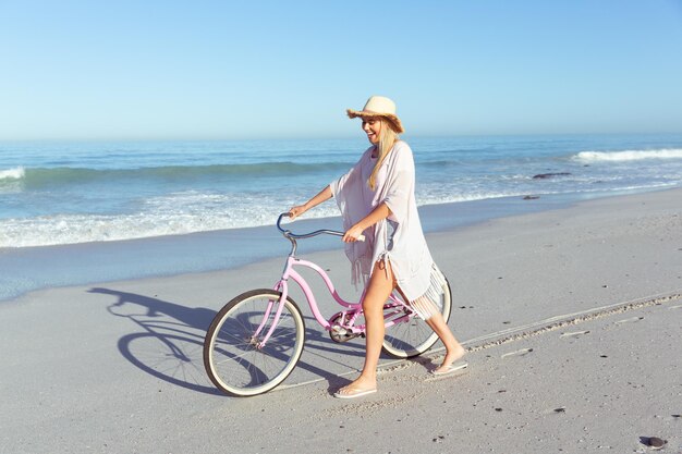 Kaukaska kobieta w kapeluszu spędzająca czas na plaży w słoneczny dzień, jeżdżąca na rowerze, z morzem w tle