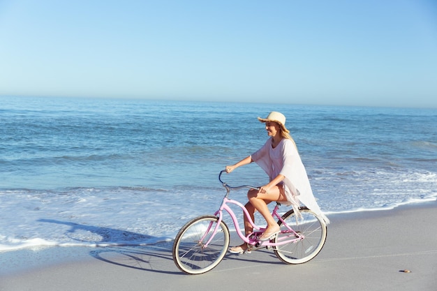 Kaukaska kobieta w kapeluszu spędzająca czas na plaży w słoneczny dzień, jeżdżąca na rowerze, z morzem w tle