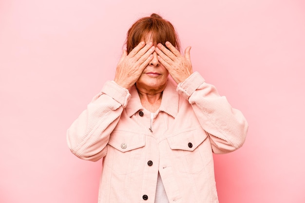 Kaukaska kobieta w średnim wieku na różowym tle boi się zasłaniać oczy rękami