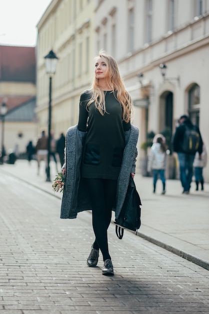 Kaukaska ładna blond młoda kobieta z długimi blond falistymi włosami chodzi na ulicy i trzyma plecaka i bukiet.