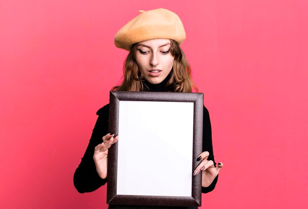 Zdjęcie kaukaska ładna blondynka maluje studenta sztuki z beretem i trzyma pustą ramkę