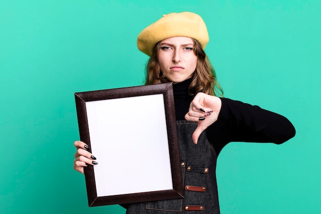Kaukaska ładna blondynka maluje studenta sztuki z beretem i trzyma pustą ramkę