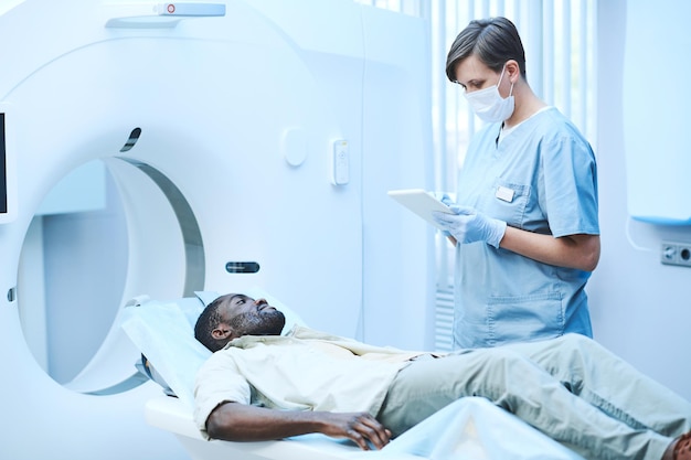 Kaukaska pielęgniarka w fartuchu i masce stojąca przy skanerze MRI i wypytująca afroamerykańską pacjentkę o objawy