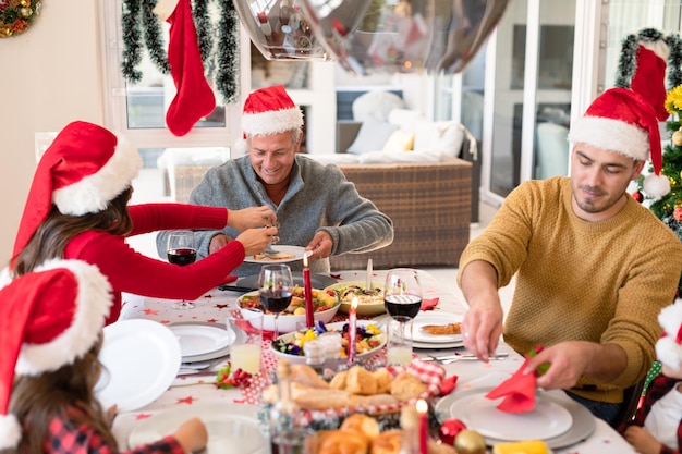 Kaukaska rodzina wielopokoleniowa w kapeluszach Mikołaja jedząca świąteczny posiłek. rodzinne święta Bożego Narodzenia i wspólne świętowanie w domu.