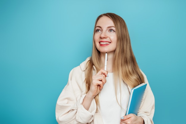 Kaukaska Studentka Myśli, Marzy, Wyobraża Sobie I Trzyma Notatnik, Notatnik, Książkę Na Białym Tle Na Niebieskiej ścianie W Studio. Testy, Egzaminy, Koncepcja Edukacji. Skopiuj Miejsce