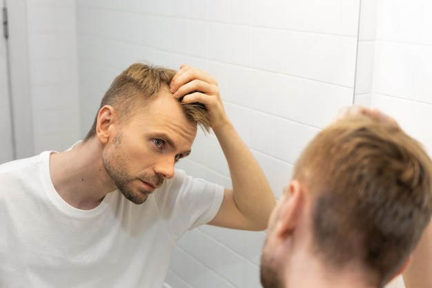 Kaukaski biały mężczyzna w średnim wieku z krótką brodą patrzy na swoje włosy w lustrze w łazience i obawia się łysienia Pojęcie problemu męskiego wypadania włosów wczesne łysienie i łysienie